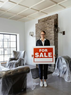 Professional real estate agent holding a 'For Sale' sign in a furnished indoor setting.